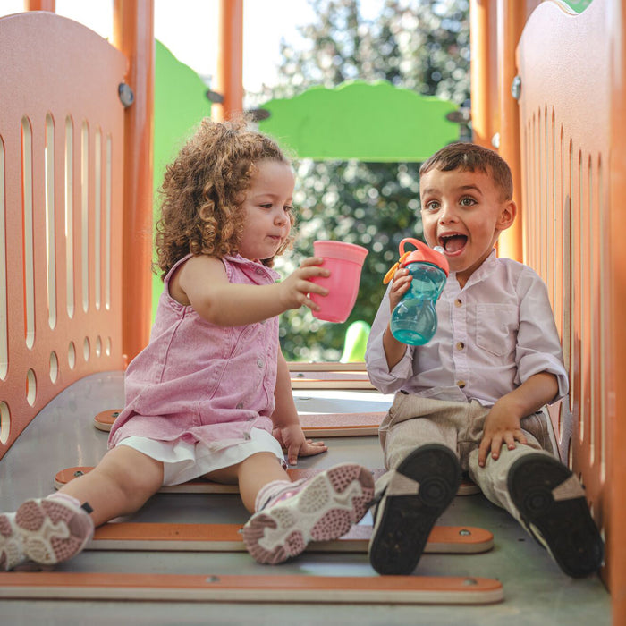 Two young children, a girl and a boy, sit together on an orange slide, smiling as they hold their Nuby 360 Wonder Cups (2 Pack - 7 oz | Pink/Aqua). Behind them, green trees complete the joyful playtime scene.