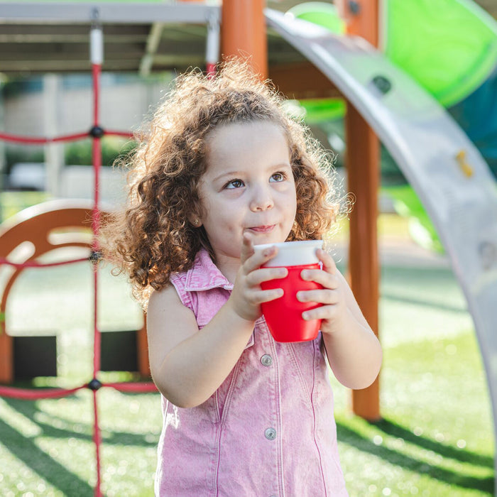 A young child with curly hair and a pink sleeveless shirt drinks outdoors from a red Nuby 360 Wonder Cup (3 Pack - 7 oz, Blue/Red/Aqua) on a playground surrounded by green grass and climbing equipment.