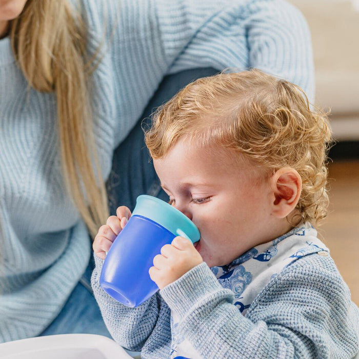 A young child with curly blonde hair, wearing a patterned bib and light blue sweater, drinks from a blue Nuby 360 Wonder Cup with Handles (2 Pack - 5 oz). An adult in a light blue sweater sits nearby.