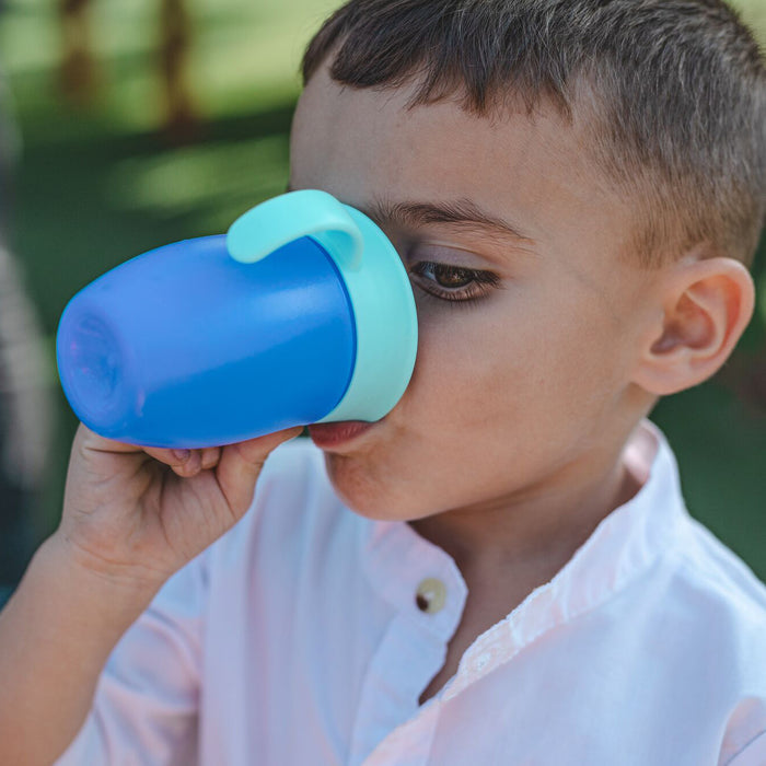 A young boy in a white shirt drinks outdoors from a blue Nuby 360 Wonder Cup with Handles (5 oz, part of the 3 Pack - Blue/Red/Aqua), with green grass in the background.