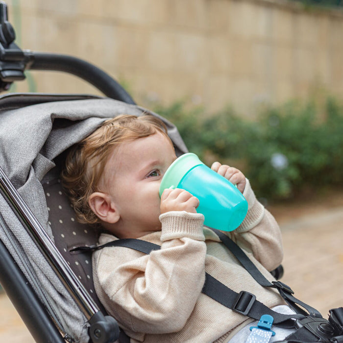 A young child in a beige sweater sips from the blue Nuby 360 Wonder Cup with Handles (3 Pack - 5 oz, Blue/Red/Aqua) while sitting in a stroller outdoors. The scene features a blurred stone wall and greenery in the background.