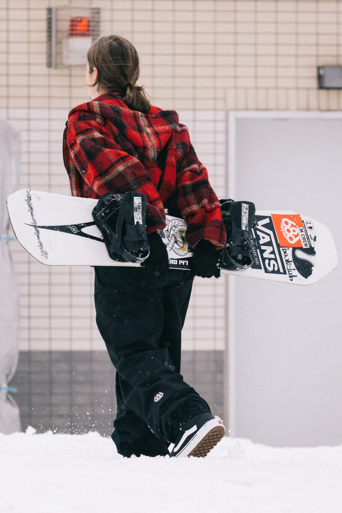 A person in a red plaid jacket and the 686 Women’s Outline™ Shell Pant walks through the snow holding a snowboard covered in stickers, facing away from the camera toward a building with tiled walls.