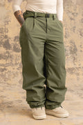 A person stands with hands in pockets against a mottled beige background, wearing the 686 Women’s Outline™ Shell Pant from 686 in olive green. She pairs the pants with white shoes and a white long-sleeve shirt, and tattoos are visible on one forearm.