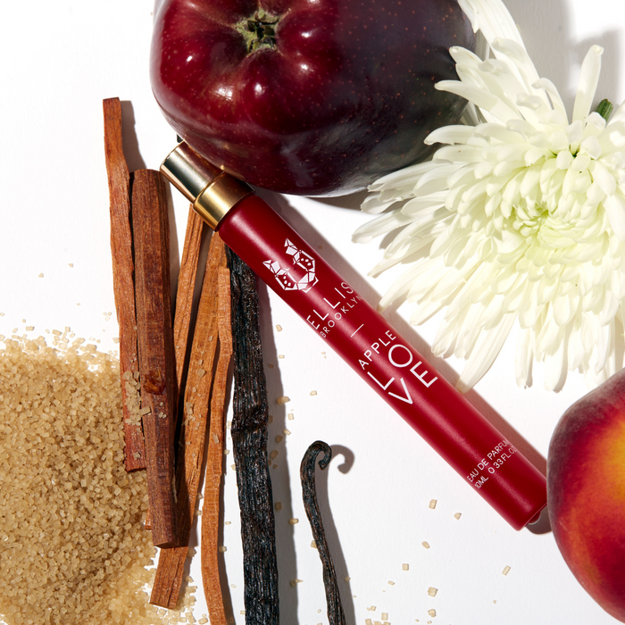 Apple Love 10ml Eau De Parfum Travel Spray next to white flower, apple, plum, sandal wood, vanilla beans, and sugar on white background.