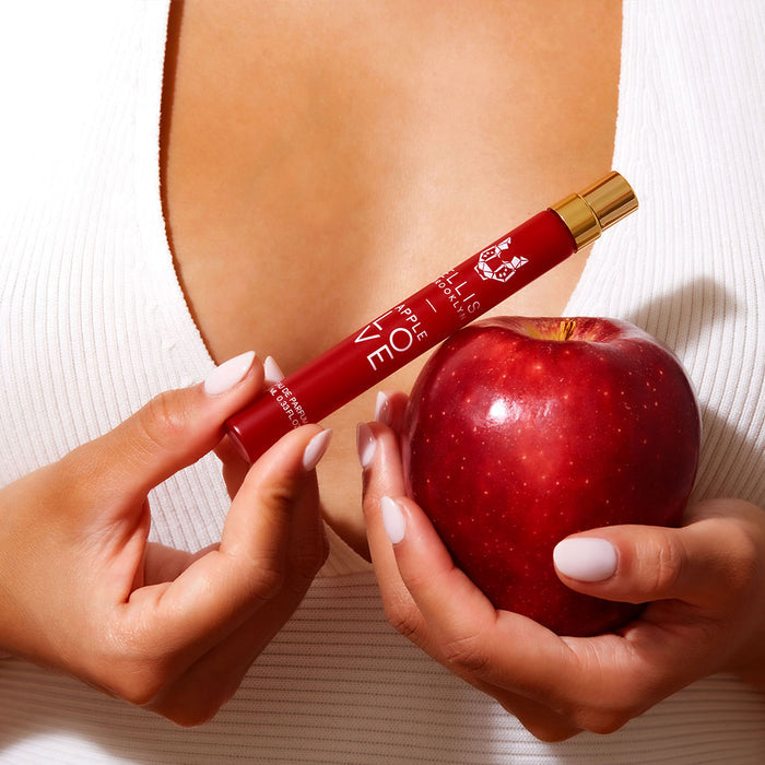 Model with light pink nail polish wearing white shirt holding Apple Love 10ml Eau De Parfum Travel Spray in one hand and an apple in the other hand, near chest.