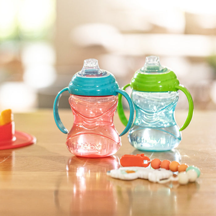 Two Nuby Clik-It Soft Spout Grip N' Sip Trainer Cups (Aqua/Grey, 2 Pack) with silicone spouts and leak-resistant design sit on a wooden table beside a baby teether and red plate in a brightly lit room.