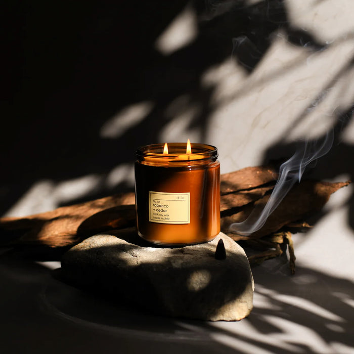 Candle in a glass jar with a label on a stone surface, surrounded by shadows of leaves.