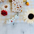 Fall Floral Bliss scrunchie phone wrist strap clipped to the matching Fall Floral Bliss Wavy Luxe iPhone case, styled on cream gingham with a white dahlia, a cream sunflower, a small orange pumpkin, and an acorn.