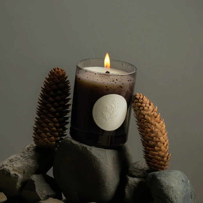 Lit Fischersund Jol holiday candle with a label on rocks and pinecones against a dark background