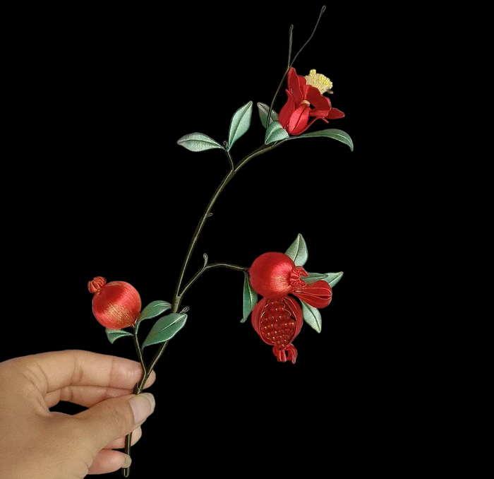Close-up of a Chanhua thread-wrapped pomegranate brooch featuring a bright red flower, a rounded fruit, and detailed green foliage.