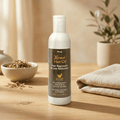 Herby Nirmal Hair Oil bottle displayed in a warm natural setting with a wooden bowl, folded towel and greenery.