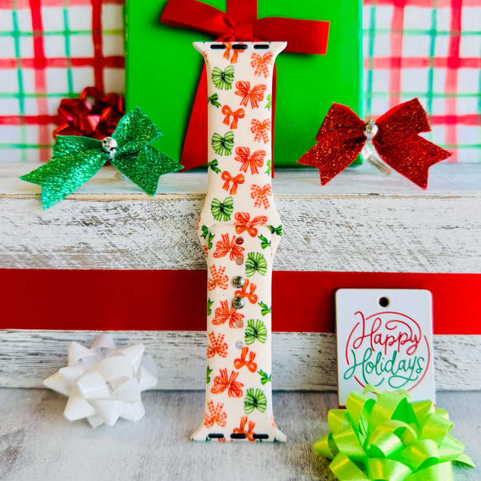 Jingle Bows Apple Watch band with red, orange, and green ribbon bows on cream background styled with wrapped Christmas presents