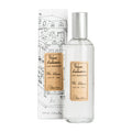 A clear glass room spray bottle with a label next to its packaging featuring a drawing of a house.