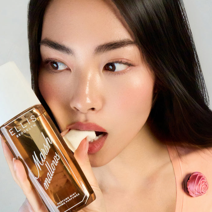 Model with brown hair wearing a peach top with a pink rose holds MARSHMALLOWS Perfume Mist 240ml against her cheek while eating a marshmallow and looking off to the side on a white background