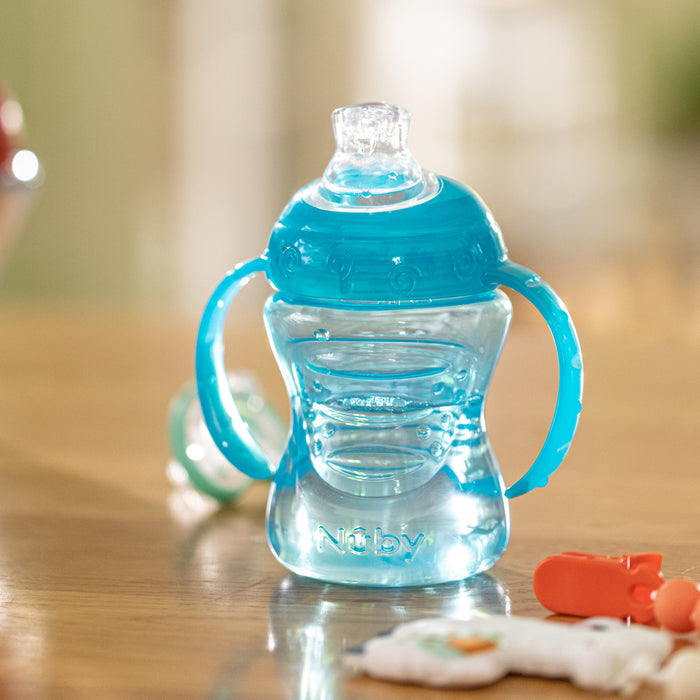 A transparent blue Nuby No-Spill Soft Spout Grip N' Sip Trainer Cup with handles and a leak-resistant silicone spout lid sits on a wooden surface, with a pacifier and toy slightly out of focus nearby.