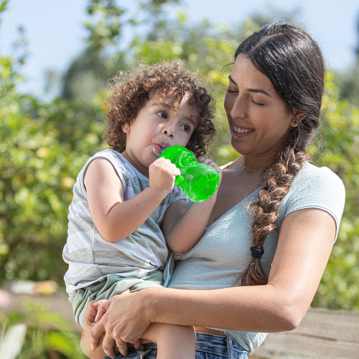 A smiling woman holds a curly-haired toddler sipping outdoors from the green, leak-resistant Nuby No-Spill Soft Spout Grip N' Sip Trainer Cup, surrounded by lush trees on a sunny day.