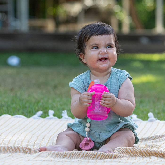 A smiling baby outdoors holds a pink Nuby No-Spill Soft Spout Grip N' Sip Trainer Cup, sitting on a yellow striped blanket in a light green outfit with a pacifier, surrounded by grass and trees.