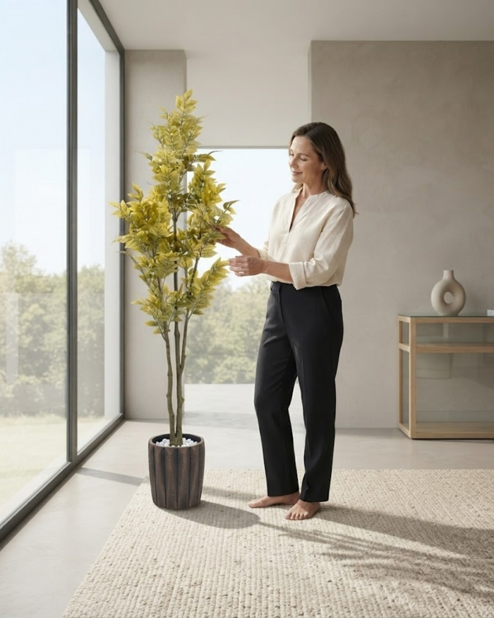 Tall artificial foliage plant in dark planter with person adjusting the stems