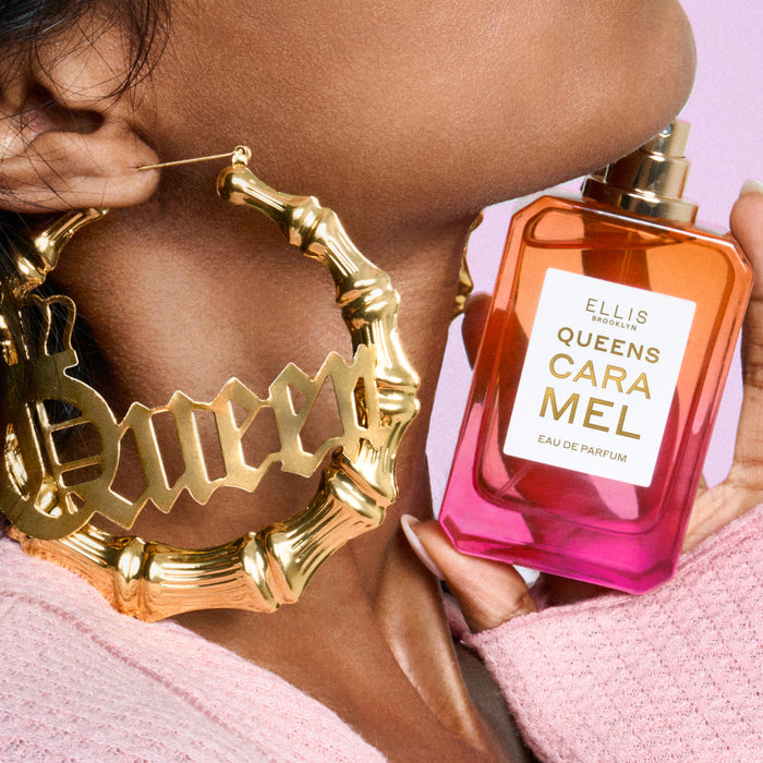 Close-up of woman wearing a pink sweater and gold hoop earrings that say "Queen" holding QUEENS CARAMEL Eau de Parfum 50ml without cap under her chin on pink background