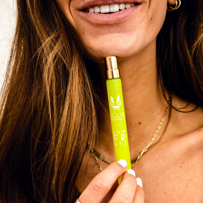 Model with brown hair and white nail polish wearing gold necklaces, earrings, and ring smiling white holding Sun Fruit 10ml Eau De Parfum Travel Spray near chin on white background.