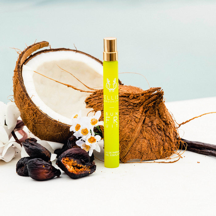Sun Fruit 10ml Eau De Parfum Travel Spray on white table in front of opened coconut, white flowers, vanilla beans, and dried figs, against pale blue background.