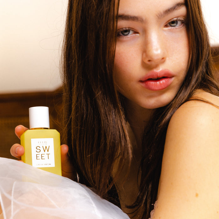 Woman with brown hair wearing a white gauze skirt holds SWEET Eau de Parfum 50ml against a brown and white background