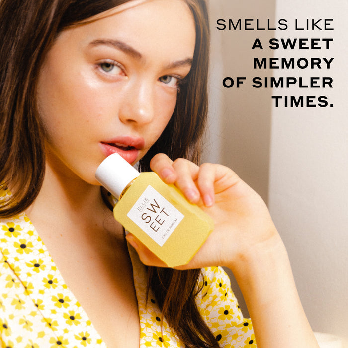 Woman with brown hair wearing a white top with yellow flowers holds SWEET Eau de Parfum 50ml against her chin against an off-white background with black text that reads "smells like a sweet memory of simpler times".