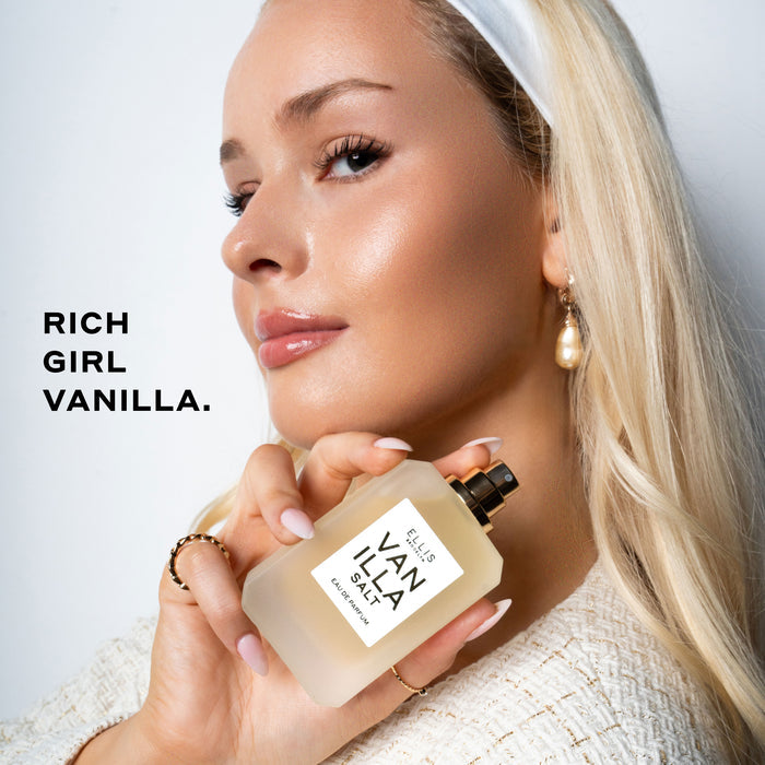 Model with blond hair and nude nail polish wearing white headband, pearl earring, gold rings, and cream boucle shirt holding Vanilla Salt 50ml Eau De Parfum without cap near shoulder on white background with black text that reads "Rich Girl Vanilla."